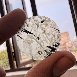 May include: A circular, translucent, clear gemstone disc featuring internal fractures and distinct black needle-like inclusions throughout the stone. The flat, polished piece has a small, centred circular hole drilled through its middle.