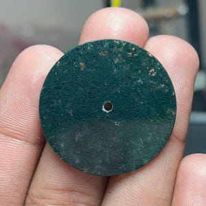 Könnte beinhalten: Ein dunkelgrünes, kreisförmiges Objekt mit einem kleinen Loch in der Mitte. Das Objekt hat eine strukturierte Oberfläche mit helleren grünen Flecken. Das Objekt wird in der Hand einer Person gehalten.