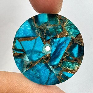 May include: A round, turquoise-colored stone pendant with a mosaic pattern. The stone features a mix of blue and green hues, with gold-colored veins running throughout. A small hole is drilled through the top for stringing.