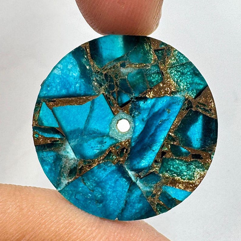 May include: A round, turquoise-colored stone pendant with a mosaic pattern. The stone features a mix of blue and green hues, with gold-colored veins running throughout. A small hole is drilled through the top for stringing.