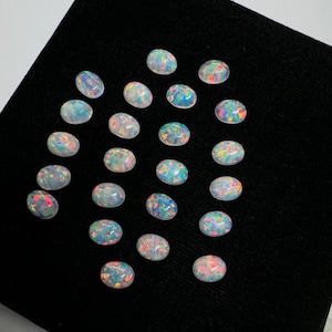 May include: A collection of 25 oval-shaped, white opal cabochons with iridescent rainbow colors. The opals are arranged in rows on a black surface.