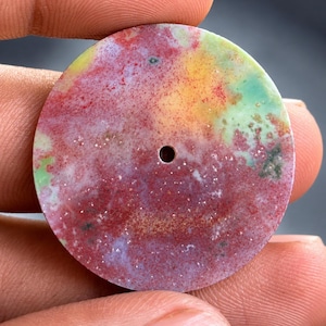 May include: A circular, flat stone pendant featuring a mottled, multi-colored surface with vibrant patches of deep red, soft yellow, and pale green. The stone has a smooth, polished texture and a small, centered circular hole for threading.