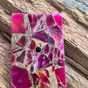 May include: A rectangular stone with a mosaic design and a central hole. The stone is a mix of pink, magenta, and gold. The irregular shapes create a unique pattern. Likely used for jewellery or crafts, the stone's dimensions are approximately 3 x 4 cm.