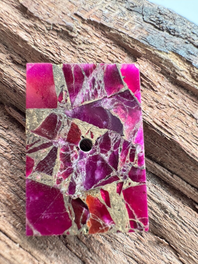 May include: A rectangular stone with a mosaic design and a central hole. The stone is a mix of pink, magenta, and gold. The irregular shapes create a unique pattern. Likely used for jewellery or crafts, the stone's dimensions are approximately 3 x 4 cm.
