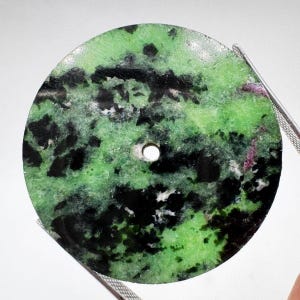 Pode incluir: Um pingente de pedra circular e plano com um padrão manchado de verde-limão vibrante, preto profundo e pequenas inclusões magenta. A peça tem uma superfície lisa e polida com um pequeno orifício circular central para suspensão.