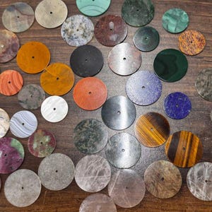 May include: A collection of various colorful, circular gemstone watch faces. The watch faces are in shades of orange, red, black, green, blue, and brown. Each face has a small hole in the center.