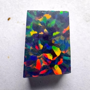 Puede incluir: Una piedra preciosa de ópalo negro rectangular con vibrantes destellos de color. La piedra muestra un mosaico de tonos rojos, amarillos, verdes y azules, creando una apariencia dinámica y llamativa. El ópalo está sobre un fondo neutro.