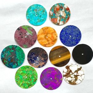 May include: A collection of eleven circular flat discs made from various stones and materials, each featuring a small centered hole. The discs display diverse patterns and colors including turquoise, blue, green, purple, orange, brown, and black.
