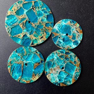 Pode incluir: Quatro discos circulares com um padrão de mosaico vibrante em turquesa e azul-petróleo com veios dourados metálicos. Cada disco tem um pequeno orifício circular centralizado. O conjunto inclui um disco grande e três discos menores.