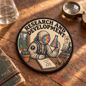 Puede incluir: Un parche bordado circular que muestra a un astronauta en un escritorio con el texto RESEARCH AND DEVELOPMENT. El astronauta sostiene un matraz colorido, escribe en un cuaderno con una estructura química y está junto a un cactus, una rana y libros.