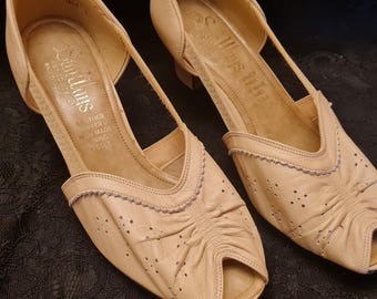 May include: A pair of beige peep-toe heeled shoes with a delicate bow detail on the front. The shoes have a light brown leather upper and a white leather lining. The brand name "Selkirk" is printed on the insole.