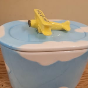 Könnte beinhalten: Keramik-Schmuckkästchen mit blauem Himmel und Wolkenmuster. Eine gelbe Flugzeugfigur sitzt auf dem Deckel. Die Box steht auf einer Holzoberfläche.
