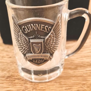 May include: Clear glass shot glass with a handle, featuring a metal emblem with the Guinness logo, wings, and the text "Traditionally Brewed 1759". The shot glass is on a wooden surface.