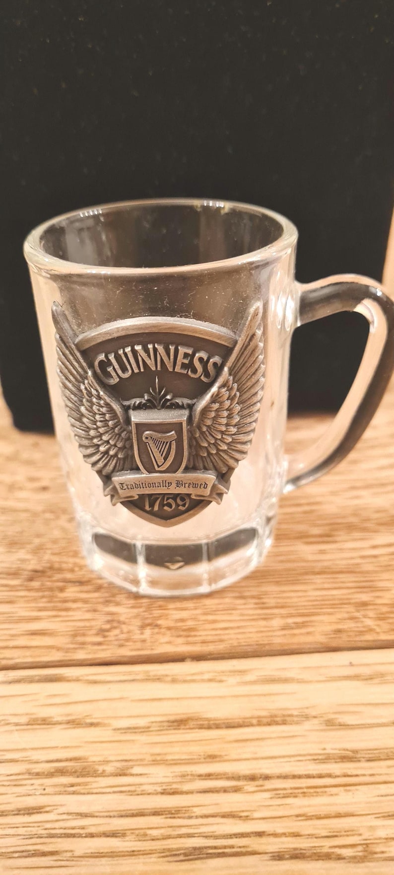 May include: Clear glass shot glass with a handle, featuring a metal emblem with the Guinness logo, wings, and the text "Traditionally Brewed 1759". The shot glass is on a wooden surface.