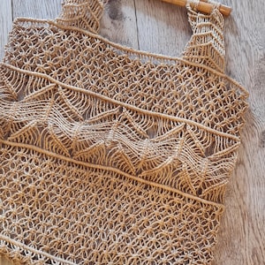 Puede incluir: Un bolso tote de macramé beige con asa de bambú. El bolso presenta un patrón tejido detallado con un diseño geométrico. El bolso está hecho de una fibra natural y tiene un estilo casual y bohemio.