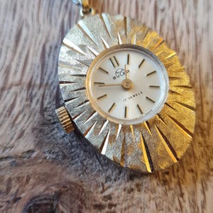 Könnte beinhalten: Eine goldfarbene Vintage-Anhängeruhr mit einem strukturierten, sonnenstrahlenförmigen Rahmen. Das Zifferblatt der Uhr ist weiß mit schwarzen Ziffern und Zeigern, und der Markenname "Buler" ist sichtbar. Die Uhr hat eine kleine Schlaufe für eine Kette.
