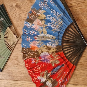 May include: Two decorative hand fans. One fan is green and yellow with a landscape design. The other fan is blue and red with a floral pattern. Both fans have black wooden frames and are open, displaying their intricate designs.