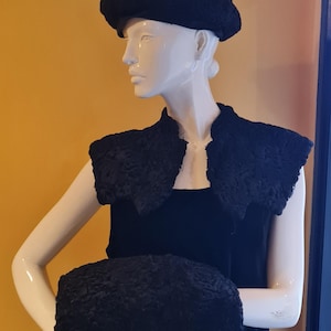 May include: A black vintage outfit displayed on a mannequin, comprising a beret, a bolero, and a muff. The set is made of a textured material, possibly fur. The mannequin is set against an orange background.