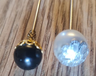 May include: Two decorative hat pins. One features a black, glossy, round bead with gold filigree. The other has a clear, round bead with a silver, glittery design. Both have long, silver metal stems.