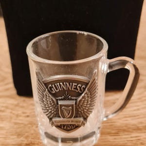 May include: Clear glass shot glass with a handle, featuring the Guinness logo. The logo includes the word "Guinness" above a harp emblem, flanked by wings, and the text "Established 1759".