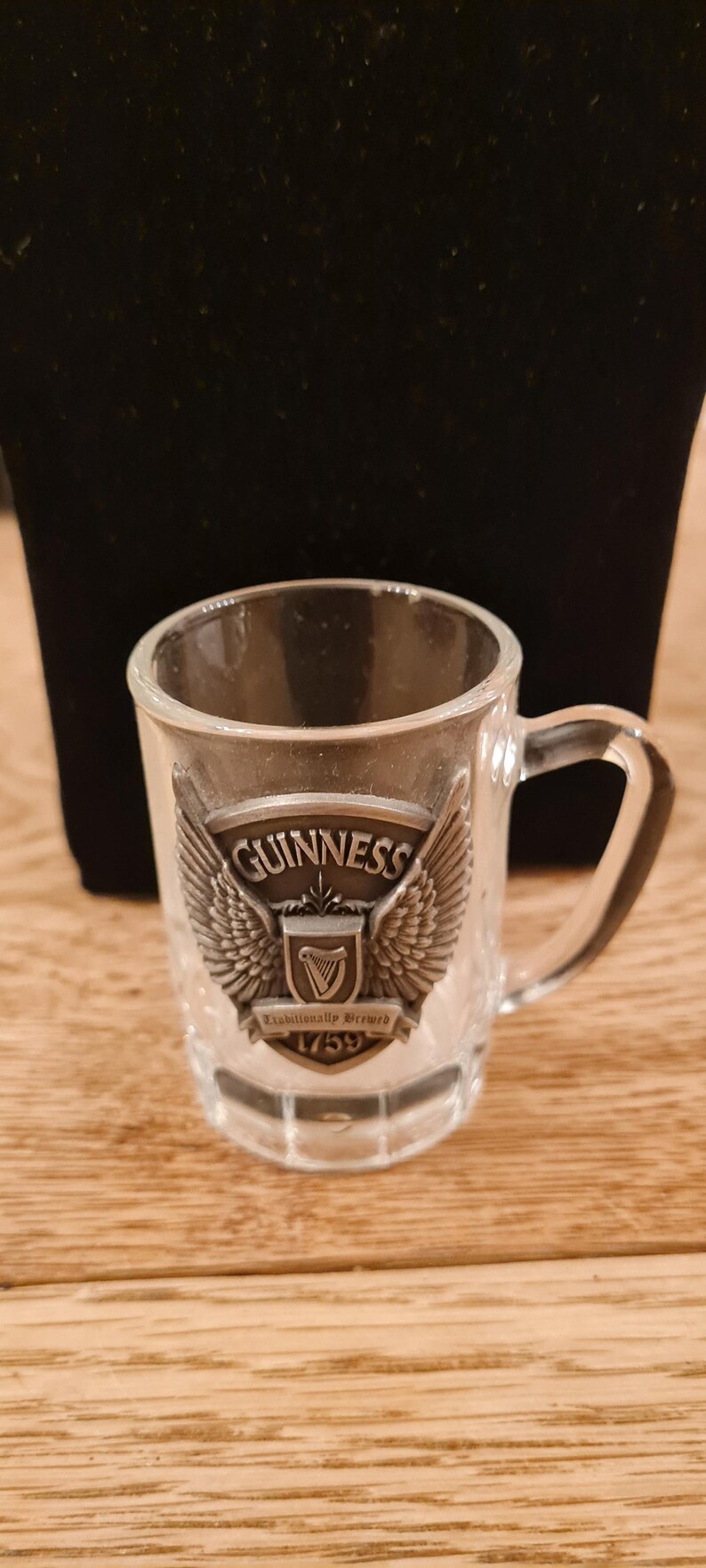 May include: Clear glass shot glass with a handle, featuring the Guinness logo. The logo includes the word "Guinness" above a harp emblem, flanked by wings, and the text "Established 1759".