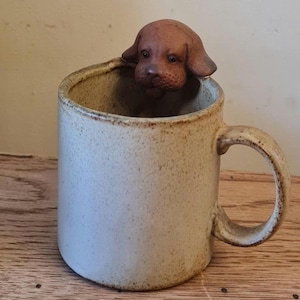 Può includere: Una tazza in ceramica beige con una statuina di cane marrone che guarda oltre il bordo. La tazza ha una consistenza macchiettata e un manico curvo. La statuina del cane è marrone con tratti del viso dettagliati. La tazza è su una superficie di legno.