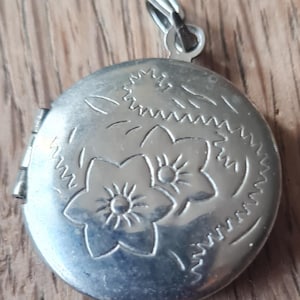 Puede incluir: Colgante de medallón redondo plateado con diseño floral. El medallón tiene una abertura con bisagras y un pequeño lazo para una cadena. El diseño floral presenta dos flores y bordes decorativos.