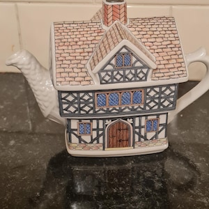 Può includere: Una teiera in ceramica bianca a forma di casa in stile Tudor. Il tetto è marrone chiaro con un motivo a mattoni e le pareti sono bianche con dettagli neri e blu. La teiera ha un camino e un beccuccio.