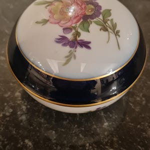 Puede incluir: Caja redonda con tapa, decorada con un motivo floral en rosa, morado y amarillo sobre fondo blanco. Tiene una banda azul marino y ribete dorado. Un recipiente de almacenamiento decorativo.