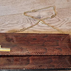 May include: Brown snakeskin-patterned clutch bag with a gold-tone chain strap. The rectangular bag has a gold-coloured clasp. The textured material of the bag contrasts with the wooden background.