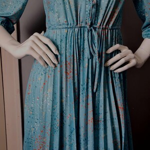 Puede incluir: Vestido vintage turquesa con cuello y mangas largas. Presenta botones delanteros, cintura ceñida con lazo y falda plisada. La tela tiene un estampado moteado en naranja y blanco. Un estilo clásico y elegante.