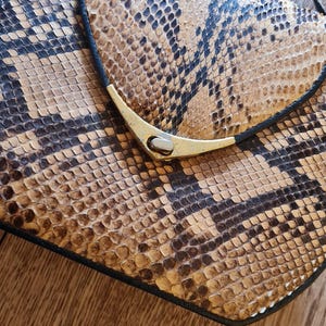Puede incluir: Bolso con estampado de piel de serpiente con cierre dorado y ribete negro. El bolso tiene una correa para el hombro y un cierre de solapa. El estampado de piel de serpiente presenta tonos marrones, beige y negros.