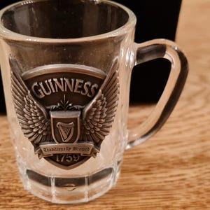 May include: A clear glass miniature beer mug with a handle. The mug features a metal emblem with the Guinness logo, including the word "Guinness", a harp, and the year 1759. The emblem is flanked by wings.