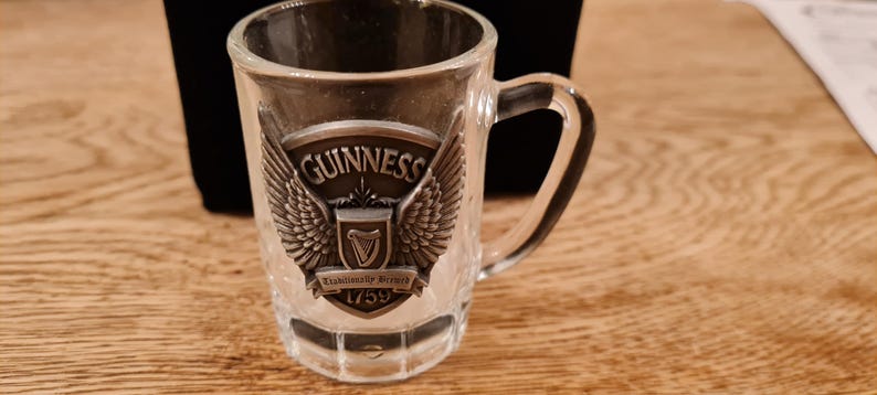 May include: A clear glass miniature beer mug with a handle. The mug features a metal emblem with the Guinness logo, including the word "Guinness", a harp, and the year 1759. The emblem is flanked by wings.