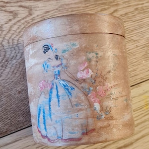 Può includere: Scatola cilindrica in legno con un disegno dipinto. L'opera raffigura una donna in abito blu con fiori rosa. La scatola ha una finitura marrone chiaro con venature del legno visibili e un coperchio.
