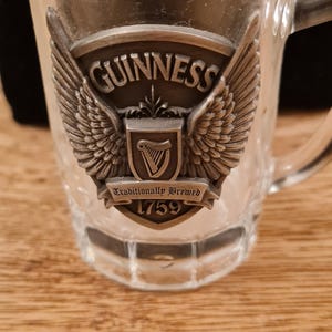May include: A clear glass mug with a handle, featuring a metal emblem with the Guinness logo, wings, and the text "Traditionally Brewed 1759." The mug is on a wooden surface.