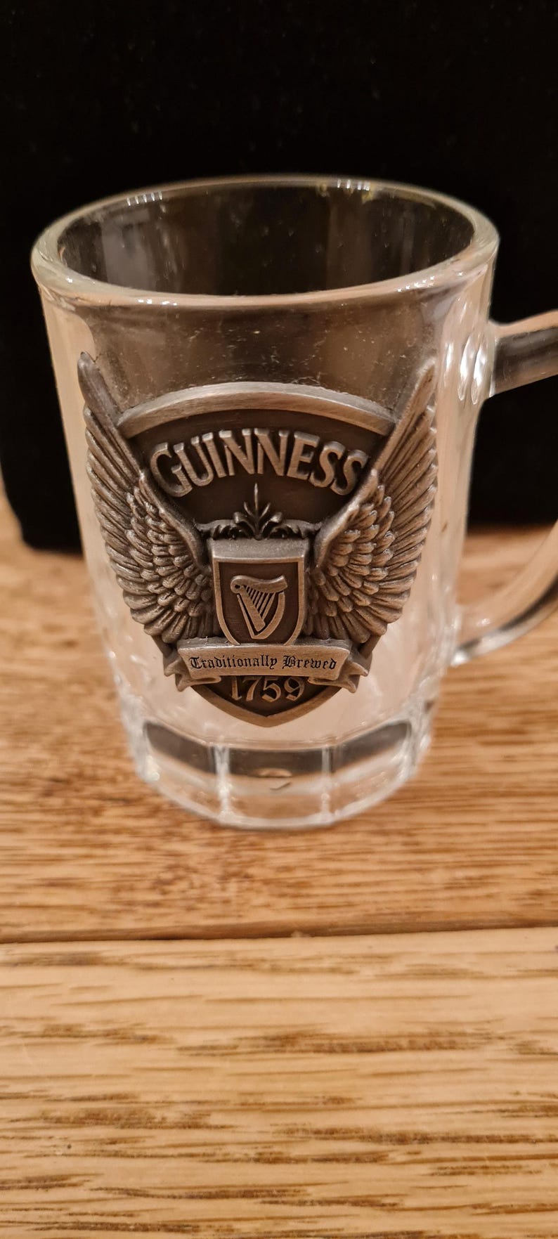 May include: A clear glass mug with a handle, featuring a metal emblem with the Guinness logo, wings, and the text "Traditionally Brewed 1759." The mug is on a wooden surface.