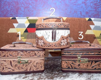 May include: Three decorative leather cases with floral and scroll designs. The cases are in shades of brown and tan, with silver-toned handles and combination locks. The center case is cylindrical with a white and brown patterned centre.