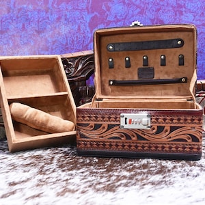 May include: Open brown leather jewelry box with a combination lock. The box has a decorative floral pattern and a tan interior with compartments for storage. A separate tray with a watch pillow is next to the open box.