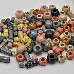Könnte beinhalten: Eine große Auswahl an Keramikperlen in verschiedenen Farben und Formen, darunter runde, ovale und zylindrische. Die Perlen sind in den Farben Rosa, Blau, Braun und Gelb.