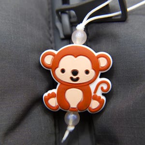 Op de afbeelding: Een bruine aapbedel met een witte omtrek en een witte staart. De bedel is bevestigd aan een wit touwtje met witte kralen.