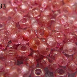 Może przedstawiać: Zbliżenie na stos małych, przezroczystych różowych koralików z połyskującym wykończeniem. Koraliki są okrągłe i mają otwór w środku. Koraliki są ułożone w losowym wzorze. #1133