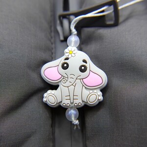 Op de afbeelding: Een grijze olifant charme met roze oren en een witte bloem op zijn hoofd. De charme is bevestigd aan een grijze rits.