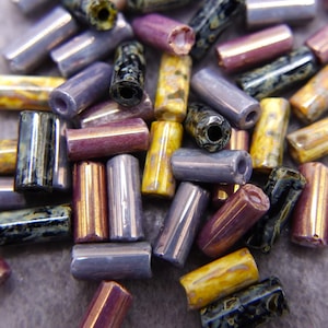 Może przedstawiać: Kolekcja iryzujących szklanych koralików w różnych kolorach, w tym fioletowym, złotym i czarnym. Koraliki mają cylindryczny kształt i gładkie, błyszczące wykończenie.