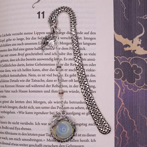 Könnte beinhalten: Silberner Drachen-Lesezeichen mit einem weißen und blauen Edelstein. Das Lesezeichen ist um ein Buch gekrümmt und hat ein detailliertes Drachen-Design mit Schuppen und Flügeln.