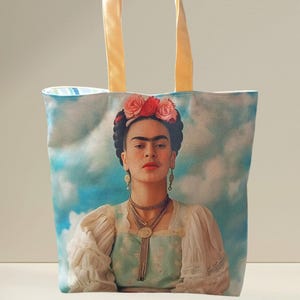 Può includere: Borsa tote con un ritratto di Frida Kahlo su uno sfondo di cielo nuvoloso. La borsa ha manici giallo chiaro e un interno a righe blu chiaro e bianco. Il ritratto mostra Kahlo con una corona di fiori, gioielli e un abito tradizionale.