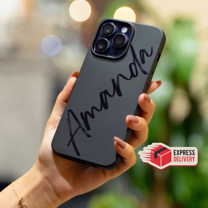 May include: Black phone case with a white inscription that says "Amanda" in a cursive font. The case is designed for a smartphone with a camera bump.