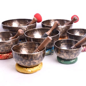 Może przedstawiać: Siedem śpiewających misek o antycznym wyglądzie z drewnianymi pałeczkami i kolorowymi poduszkami. Miski są wykonane z metalu i mają młotkowane wykończenie.