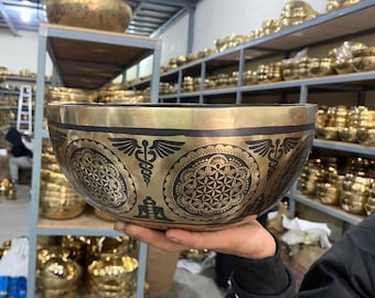 May include: A large, golden singing bowl with a black and silver design. The bowl has a flower of life pattern, a caduceus symbol, and a figure in a meditative pose. The bowl is sitting on a shelf with other singing bowls.
