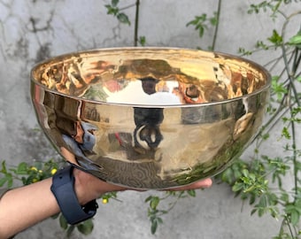 May include: A large, golden singing bowl with a hammered surface. The bowl is being held in a person's hand.